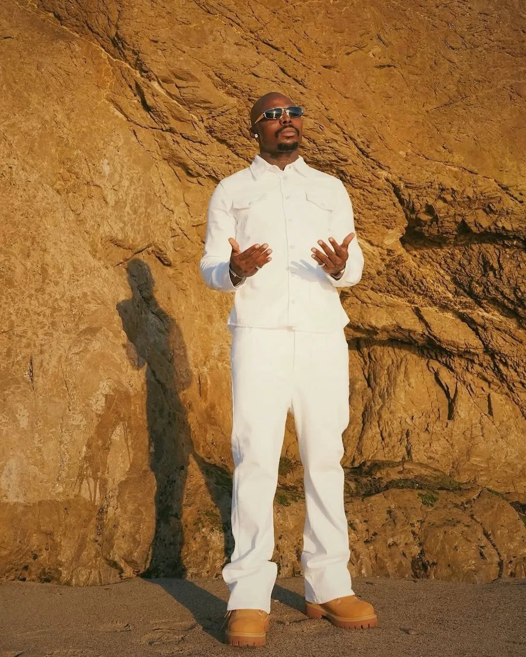 Man in all white outfit with tan boots posing against a rock wall.