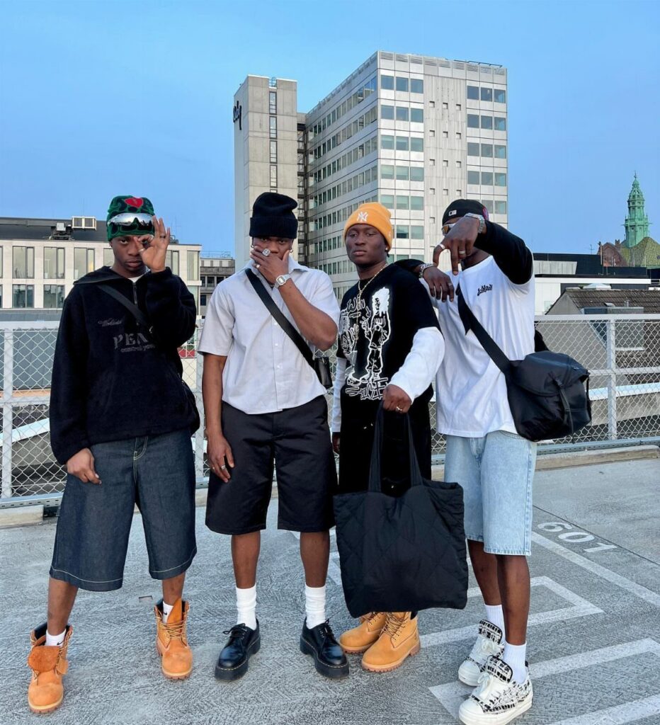 Four men posing on a rooftop wearing streetwear and knee-length bermuda shorts.