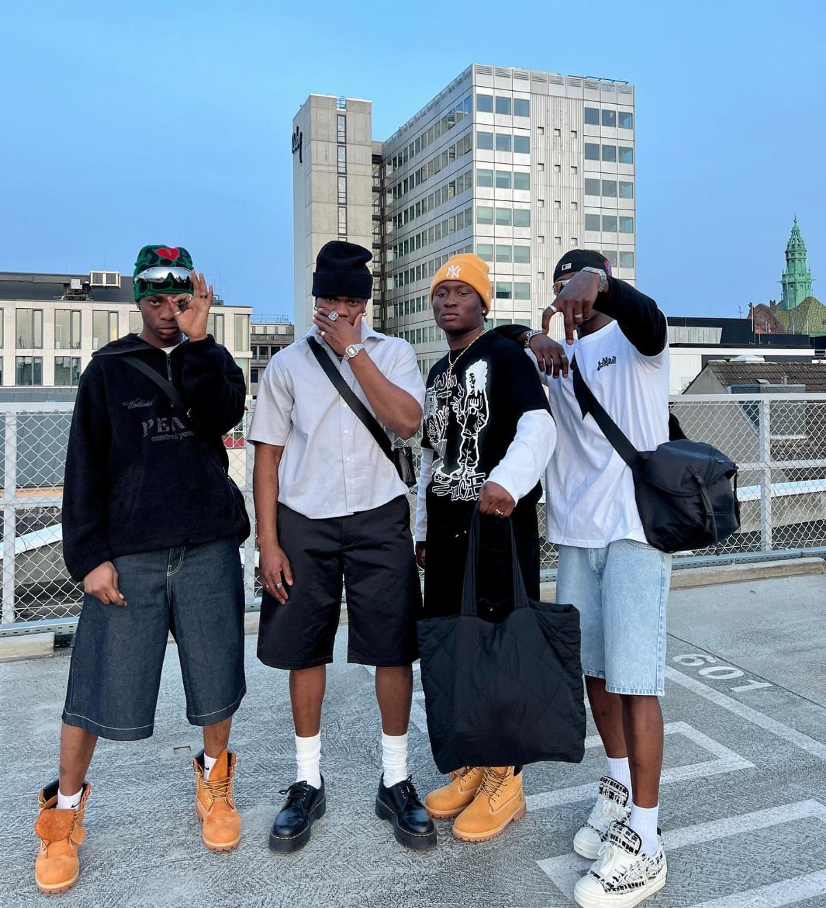 Four men posing on a rooftop wearing streetwear and knee-length bermuda shorts.