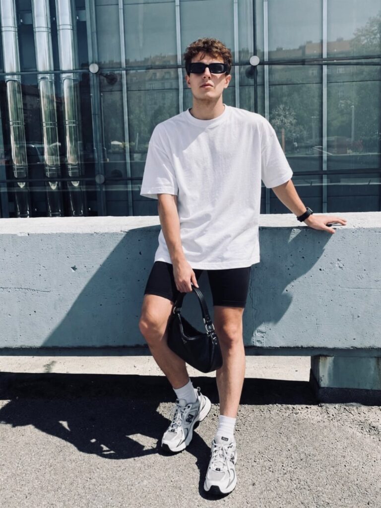 Man in white oversized t-shirt and black biker shorts leaning on concrete ledge
