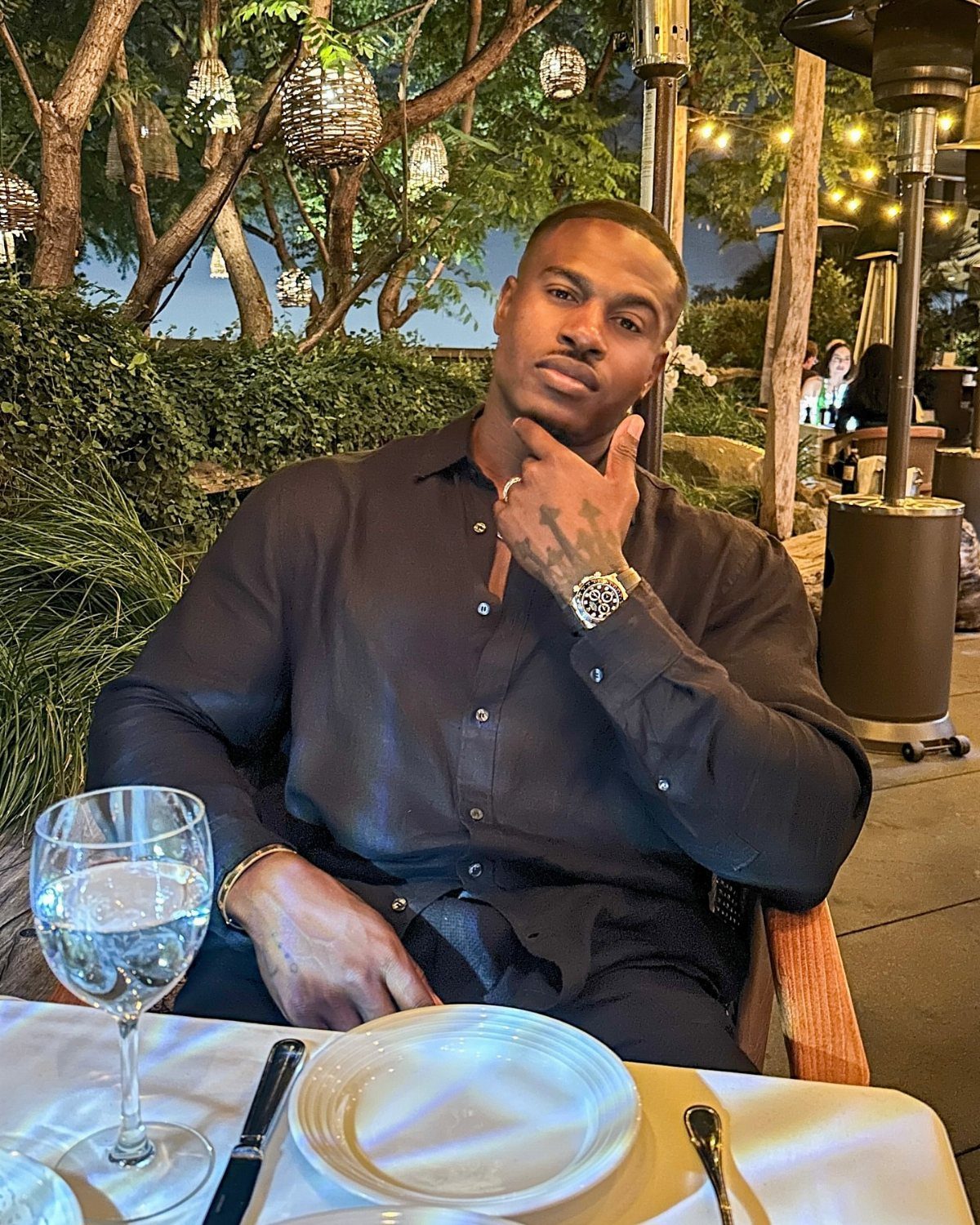 Man in black button-down shirt and trousers sitting at an outdoor dinner table.