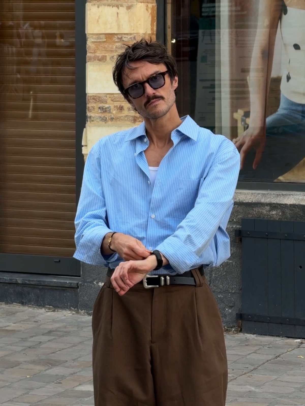 Man wearing a light blue striped shirt and brown high-waisted trousers with sunglasses.