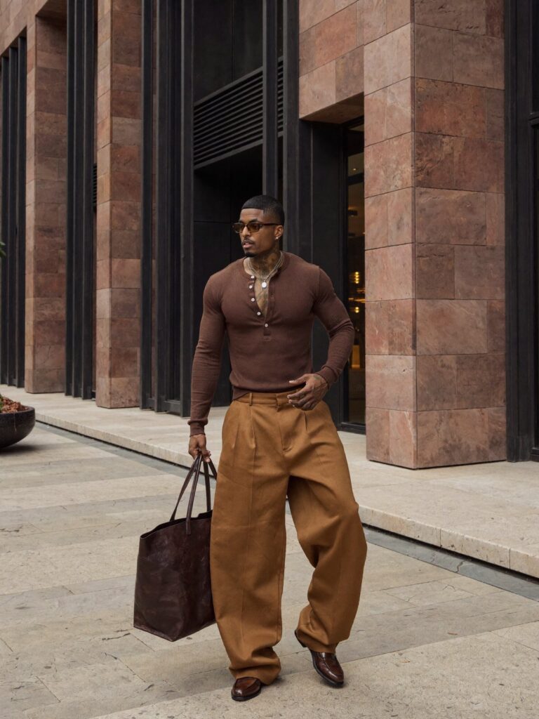 Man wearing a brown henley shirt and wide-leg brown trousers with a leather tote.