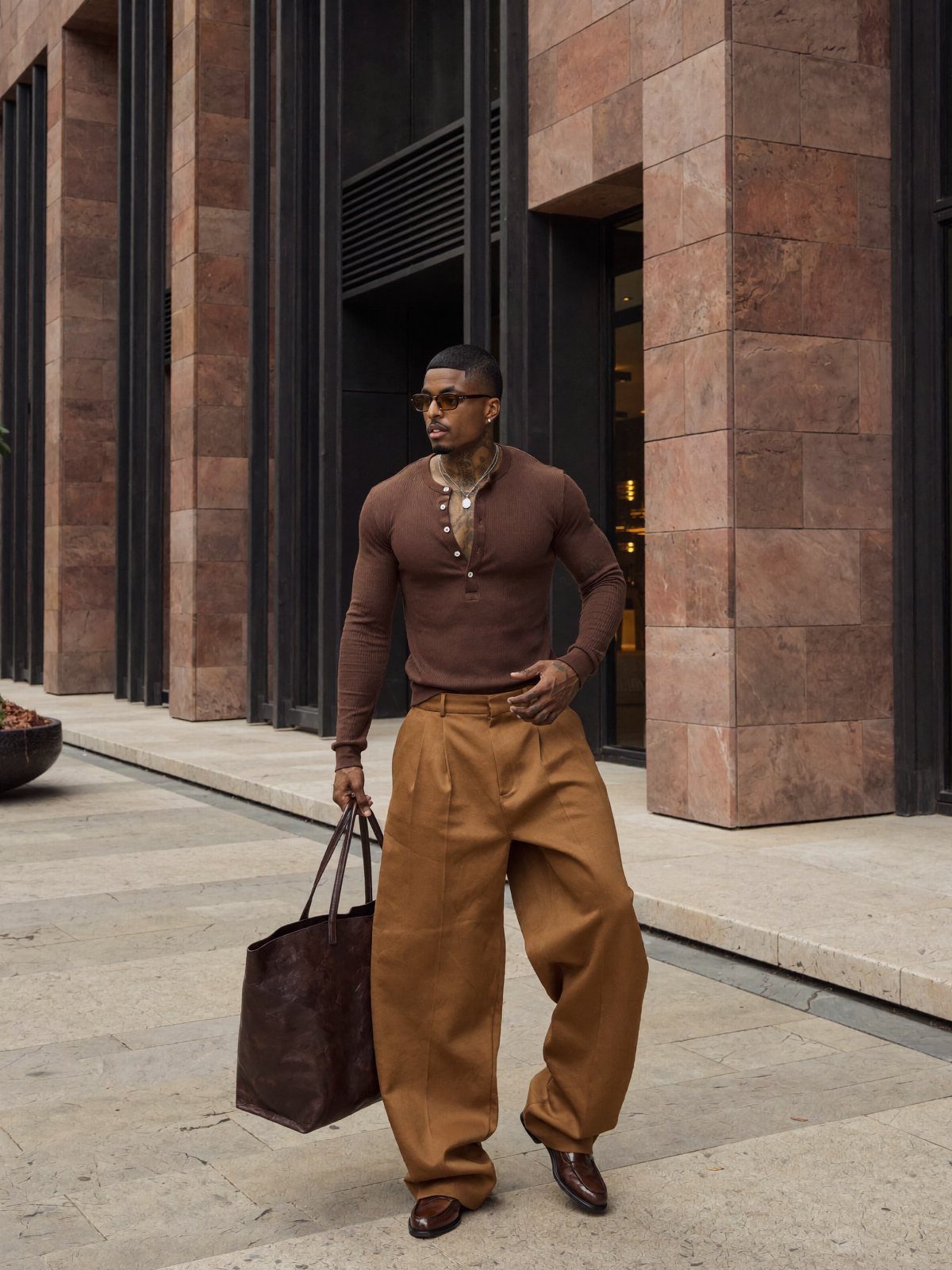 Man wearing a brown henley shirt and wide-leg brown trousers with a leather tote.