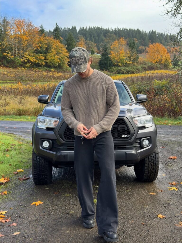 Man wearing a brown knit sweater and black trousers standing before a black truck.