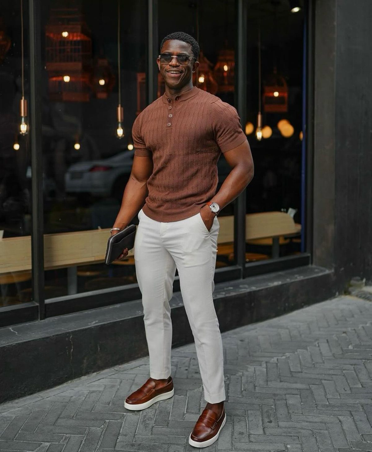 Man wearing a brown henley shirt and white trousers standing on a sidewalk.