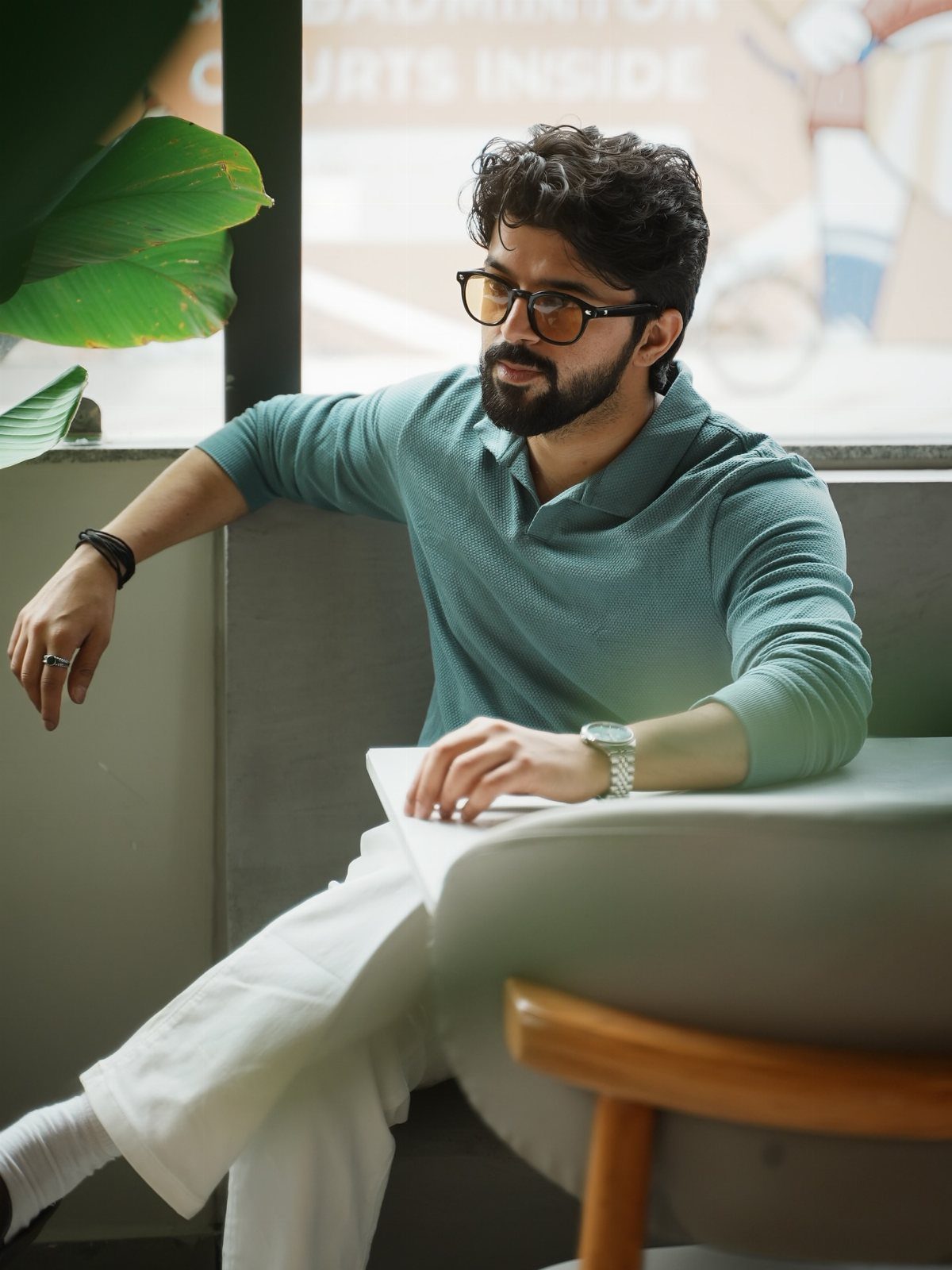 Man wearing a teal long-sleeved polo shirt and white trousers sitting indoors.