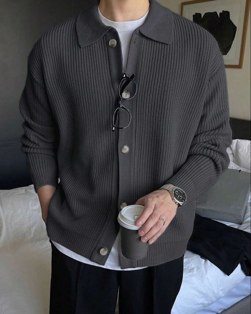 Person wearing a ribbed gray cardigan, white t-shirt, and black trousers holding coffee.