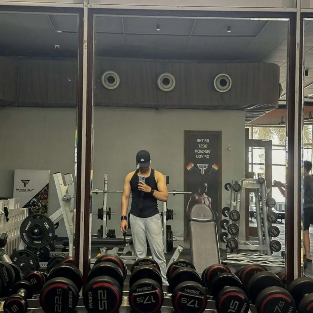 Man in gym attire taking a mirror selfie surrounded by dumbbells and equipment