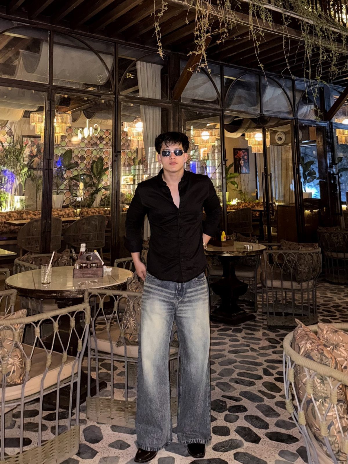 Man in black button-down shirt and wide-leg blue jeans standing in a patio.