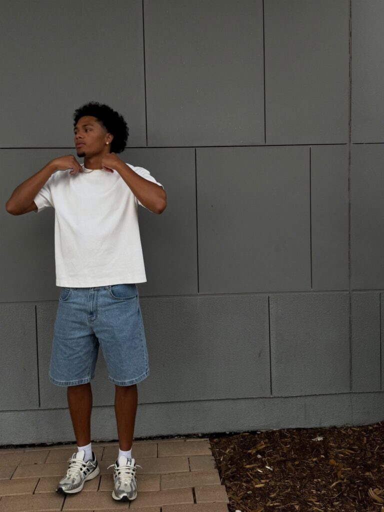 Man wearing a white t-shirt and blue denim shorts against a grey wall.