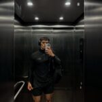 Man in black athletic wear taking a mirror selfie inside a metallic elevator.