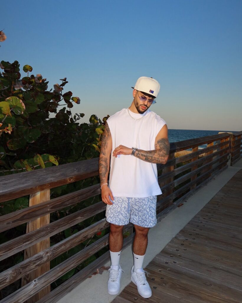 Man wearing white sleeveless shirt and patterned blue shorts on a wooden boardwalk.