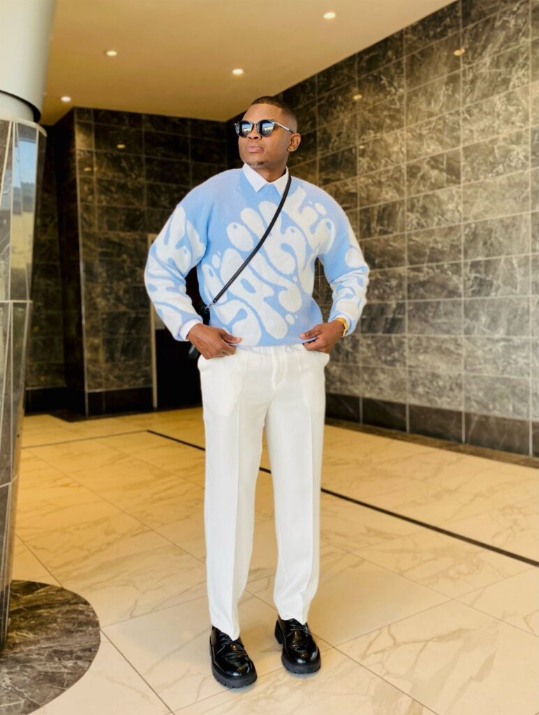 Man wearing a blue patterned sweater, white pants, and black shoes in a lobby