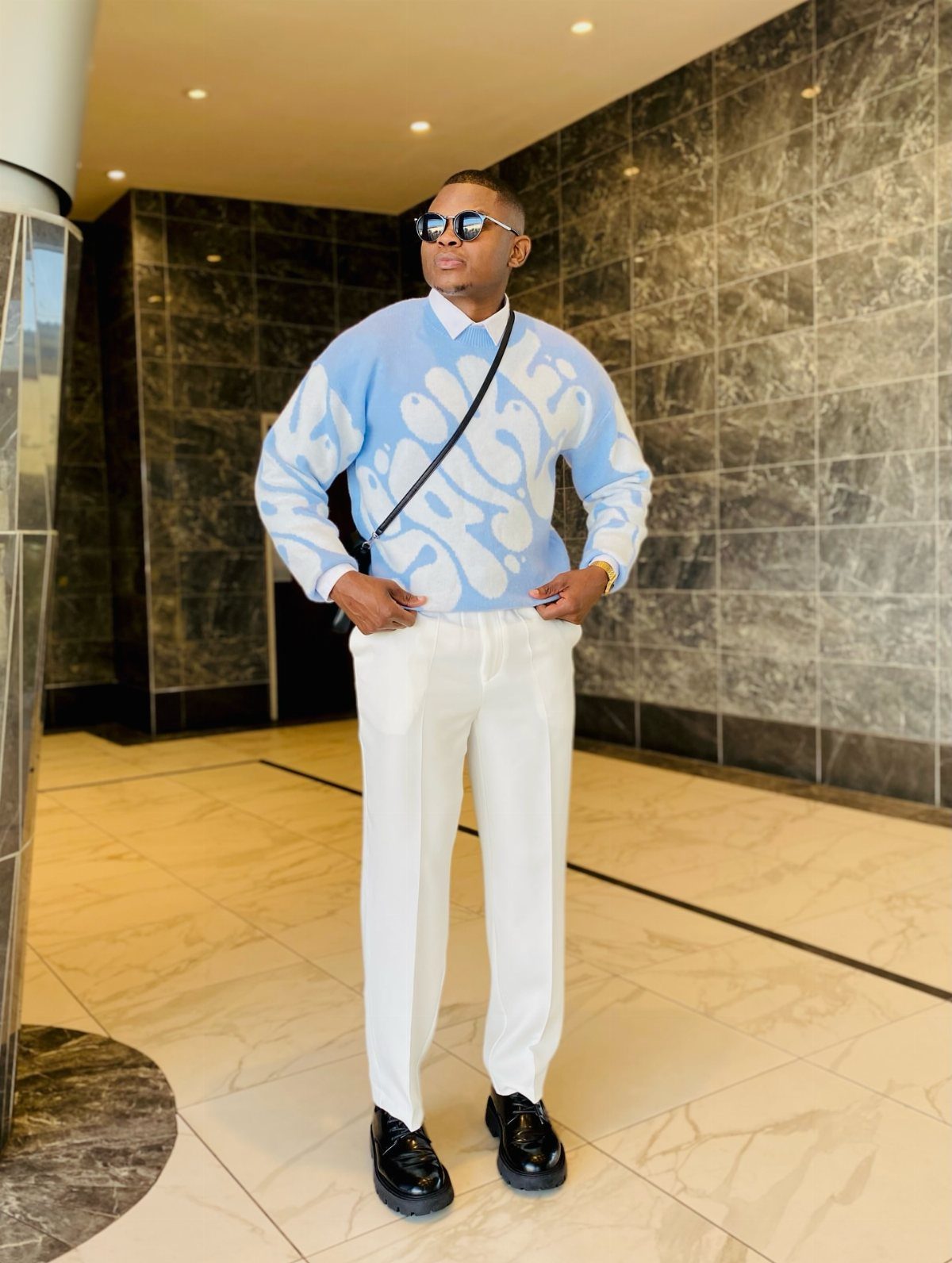 Man wearing a blue patterned sweater, white pants, and black shoes in a lobby