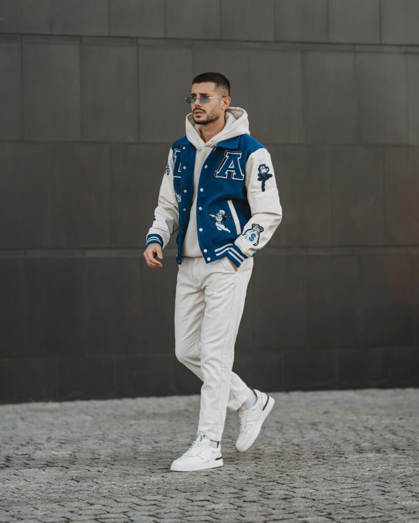 Man wearing a blue and white varsity jacket, white hoodie, and white trousers.