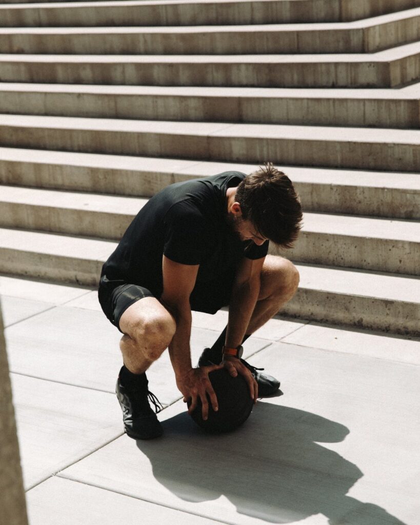 Man in black athletic wear crouching with a medicine ball by concrete stairs.
