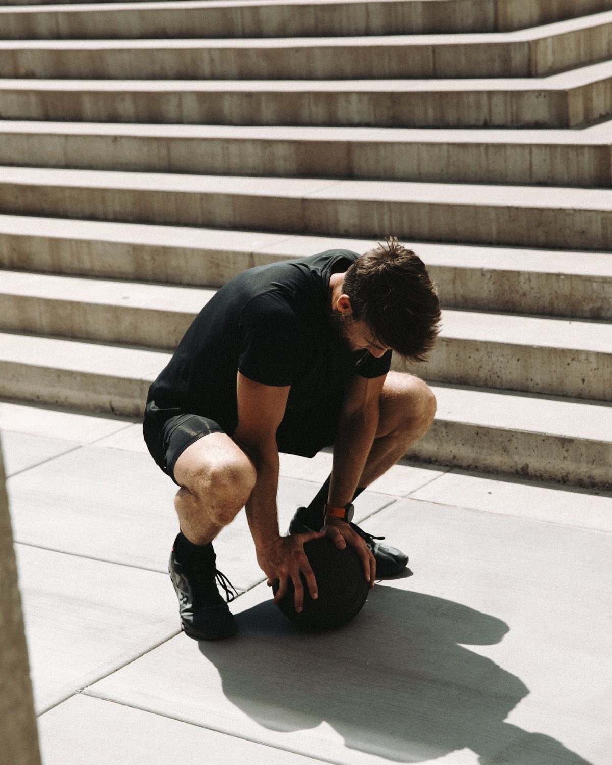 Man in black athletic wear crouching with a medicine ball by concrete stairs.