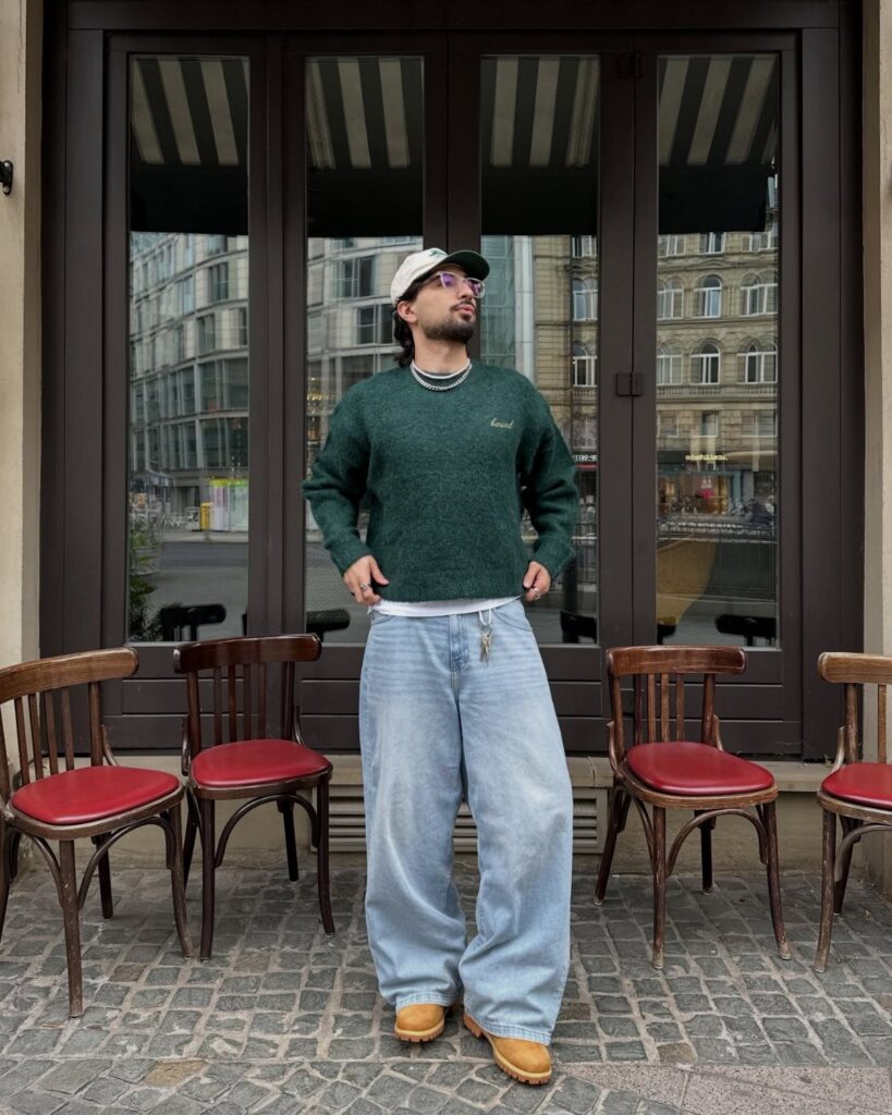 Man wearing a dark green sweater and wide-leg light blue jeans standing outdoors.
