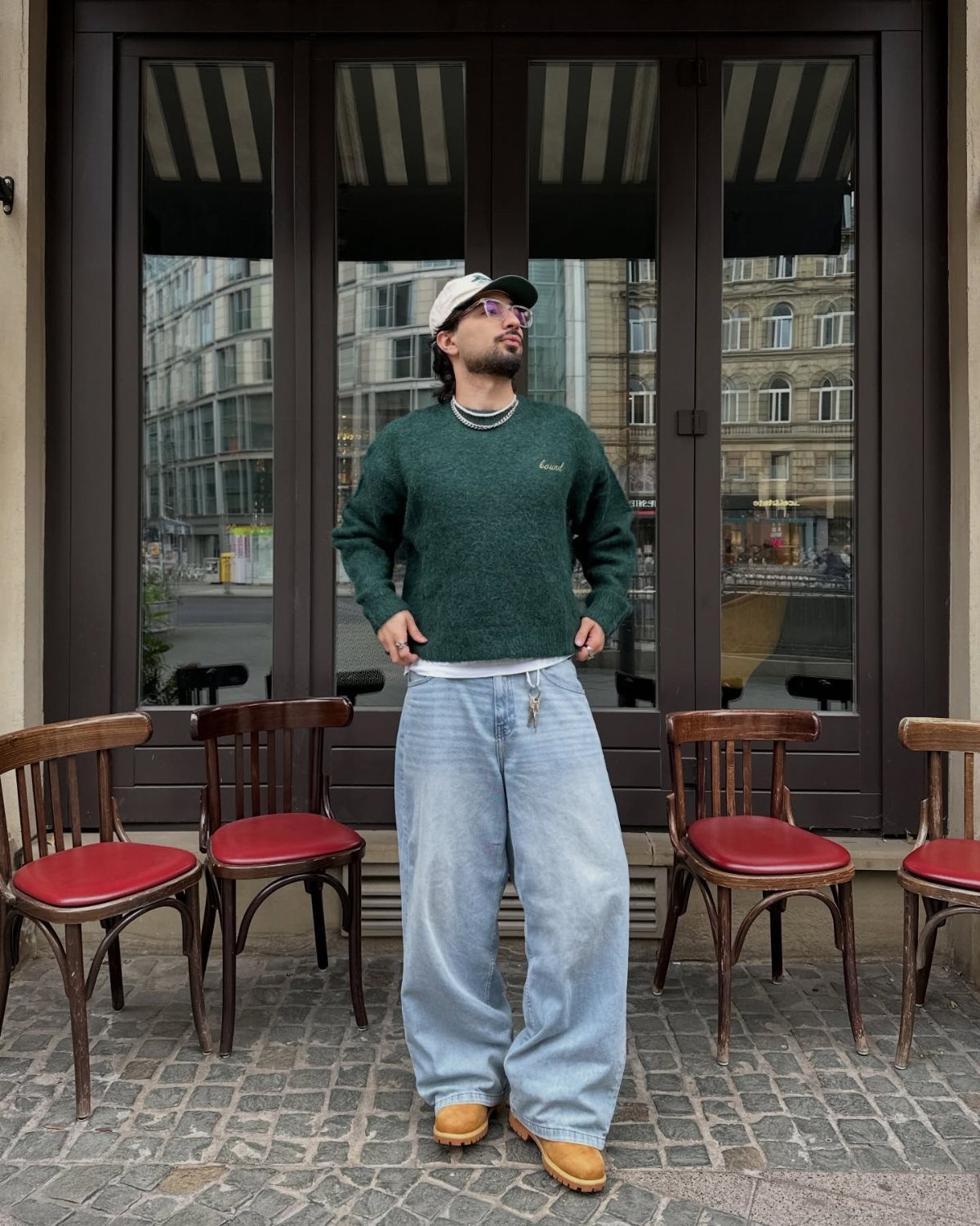 Man wearing a dark green sweater and wide-leg light blue jeans standing outdoors.